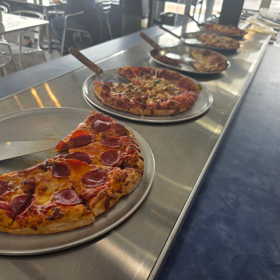 Multiple pizzas with various toppings, including pepperoni, on metal trays in a restaurant setting.