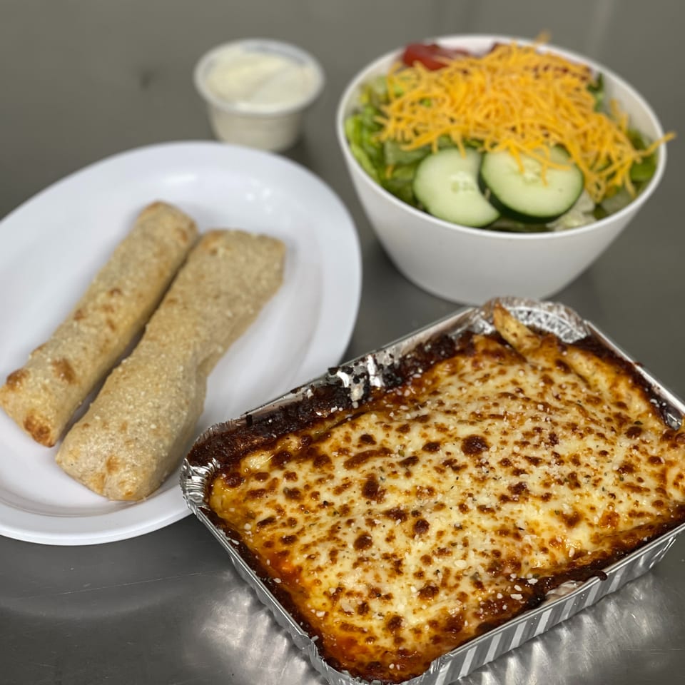 Baked lasagna in a foil pan, breadsticks, and a side salad with dressing on a metal surface.