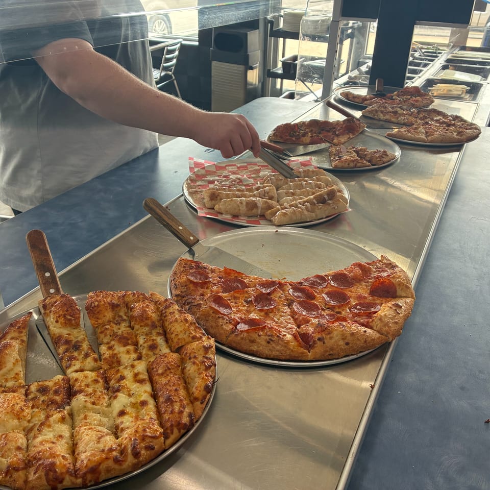 A person serving cinnamon rolls from a buffet line with various pizzas and breadsticks.